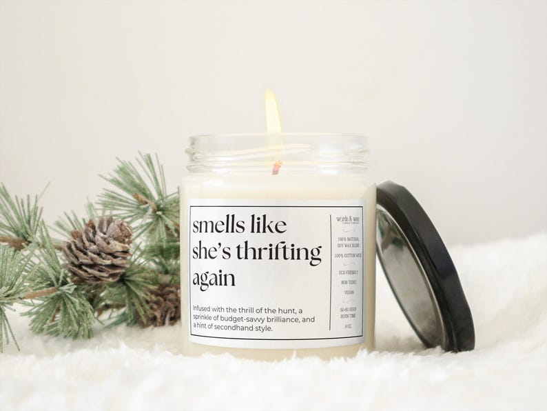 May include: A white candle in a glass jar with a black label that reads "smells like she's thrifting again". The label also includes the text "Infused with the thrill of the hunt, a sprinkle of budget-savvy brilliance, and a hint of secondhand style."