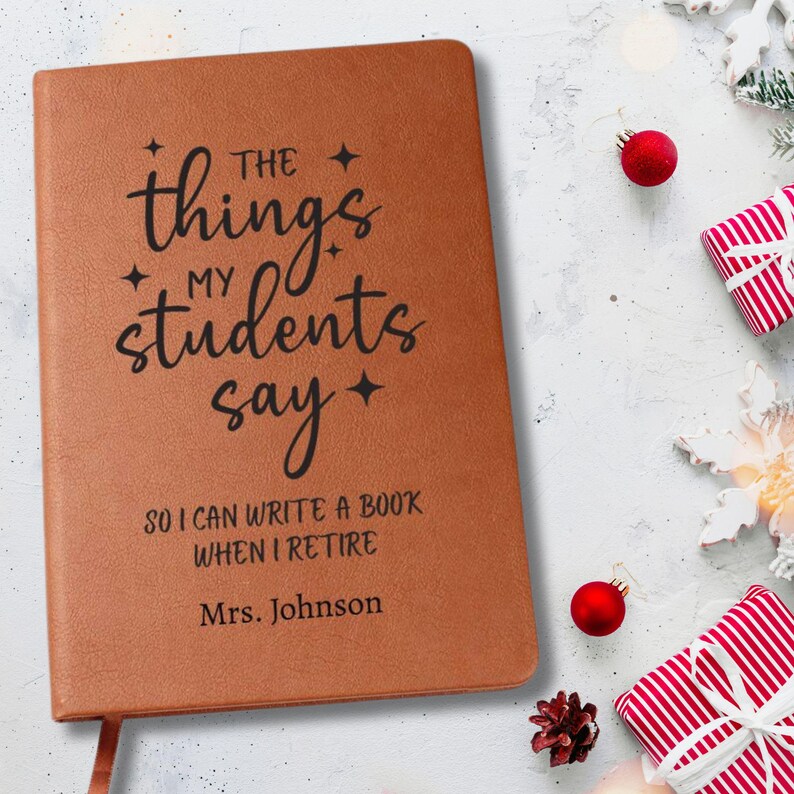 May include: A brown leather journal with the text "The things my students say" in black script. The journal also includes the text "So I can write a book when I retire" and "Mrs. Johnson". The journal is on a white surface with Christmas decorations.