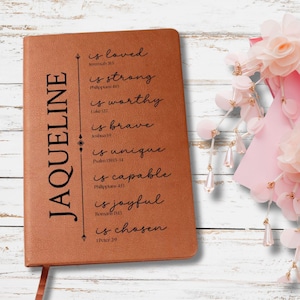 May include: A brown leatherette journal with the name "JAQUELINE" in large black letters on the left side. The journal has inspirational text on the right side, including "is loved" and "is strong". A pink ribbon and floral arrangement are in the background.