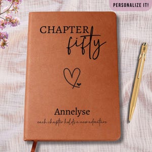 May include: A brown leather journal with the words "CHAPTER fifty" and the name "Annelise" printed on the cover. A heart graphic is below the words "fifty". The text "each chapter holds a new adventure" is printed at the bottom. A gold pen rests on the right.