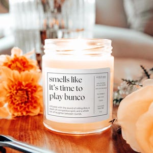 May include: A clear glass candle jar with a white label that reads "smells like it's time to play bunco". The candle is lit and the wax is a light yellow color. The candle is surrounded by orange and yellow flowers.