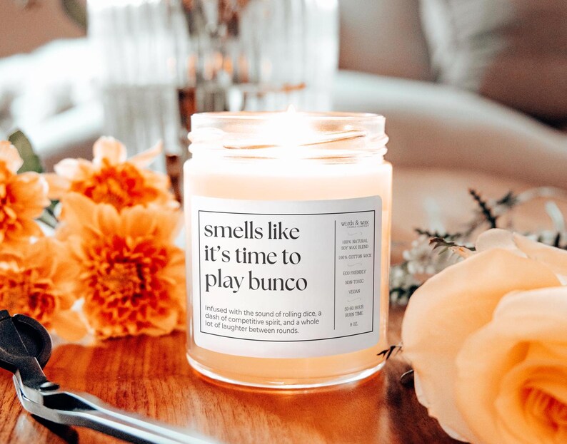 May include: A clear glass candle jar with a white label that reads "smells like it's time to play bunco". The candle is lit and the wax is a light yellow color. The candle is surrounded by orange and yellow flowers.
