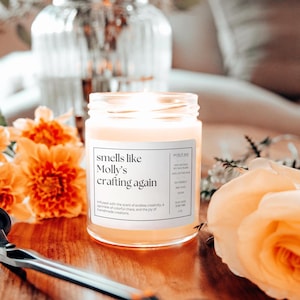 May include: A clear glass candle jar with a white label that reads "smells like Molly's crafting again". The candle is surrounded by orange and yellow flowers.