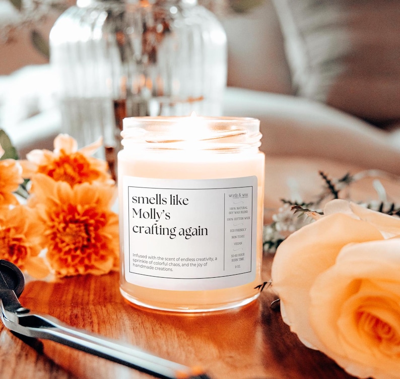 May include: A clear glass candle jar with a white label that reads "smells like Molly's crafting again". The candle is surrounded by orange and yellow flowers.