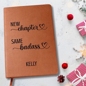 May include: A brown leather journal with the text "NEW chapter", "SAME badass", and the name "KELLY" in black. The journal has a ribbon bookmark. The background includes Christmas ornaments and wrapped gifts.