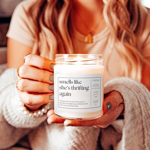 May include: A white candle jar with a label that reads "smells like she's thrifting again". The label also includes a description of the scent, which is infused with the thrill of the hunt, a sprinkle of budget-savvy brilliance, and a hint of secondhand style.