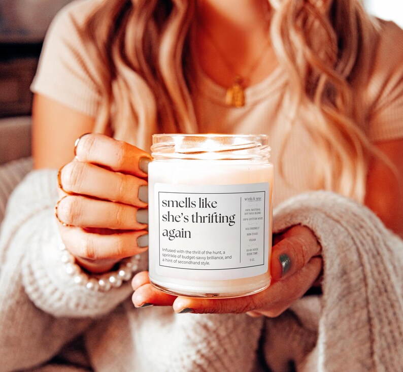 May include: A white candle jar with a label that reads "smells like she's thrifting again". The label also includes a description of the scent, which is infused with the thrill of the hunt, a sprinkle of budget-savvy brilliance, and a hint of secondhand style.