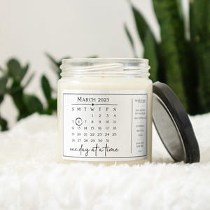 May include: A white candle with a black and white label that says "MARCH 2023" and has a calendar printed on it. The label also says "one day at a time".