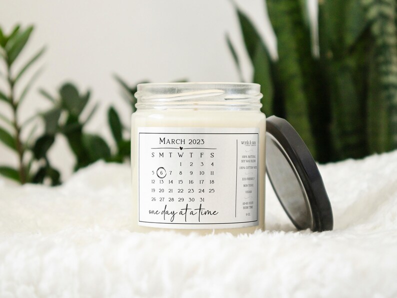May include: A white candle with a black and white label that says "MARCH 2023" and has a calendar printed on it. The label also says "one day at a time".