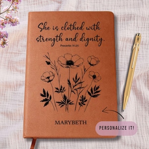 May include: A brown leather journal with the text "She is clothed with strength and dignity" and "Proverbs 31:25". The journal features a floral design and the name "MARYBETH". A gold pen rests beside the journal. The words "PERSONALIZE IT!" are also visible.