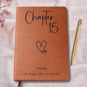 May include: A brown leather journal with the words "Chapter 15" and a heart symbol. The name "Natalia" and the phrase "each chapter holds a new adventure" are also printed on the journal. A gold pen rests beside the journal.