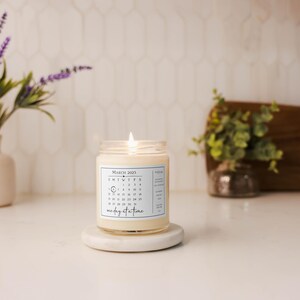 May include: A white candle with a calendar design on the label. The candle is labeled "March 2021" and has the number "10" circled. The text "one day at a time" is printed below the calendar. The candle is sitting on a white pedestal.