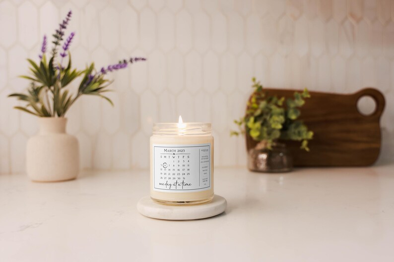 May include: A white candle with a calendar design on the label. The candle is labeled "March 2021" and has the number "10" circled. The text "one day at a time" is printed below the calendar. The candle is sitting on a white pedestal.