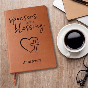 以下が含まれることがあります： 「sponsors are a blessing」の文字と、ハートと十字架のデザインが施された茶色の革製ジャーナル。ジャーナルは「Aunt Jenny」の名前でパーソナライズされています。コーヒーカップ、メガネ、ラップトップが木製の机の上にあります。