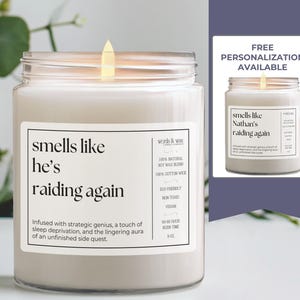 May include: A white candle with a glass jar and a black label that reads "smells like he's raiding again". The label also includes the text "Infused with strategic genius, a touch of sleep deprivation, and the lingering aura of an unfinished side quest."