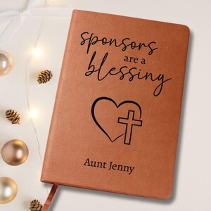 以下が含まれることがあります： 「Sponsors are a blessing」の文字と、ハートと十字架のデザインが施された茶色の革製ジャーナル。下部には「Aunt Jenny」の名前が印刷されています。ジャーナルは、クリスマスの飾りとともに白い表面に置かれています。