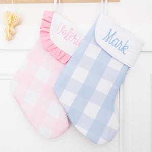 May include: Two Christmas stockings with white cuffs, one featuring a pink gingham pattern with a ruffled edge and the name Valerie embroidered in pink, and the other featuring a light blue gingham pattern with the name Mark embroidered in blue.