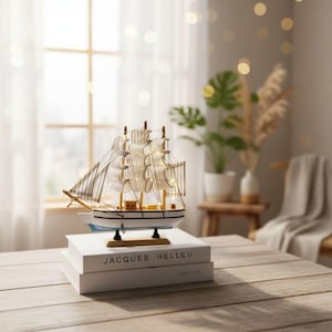Könnte beinhalten: Ein dekoratives Modellsegelschiff mit weißen Segeln und einem weißen Rumpf, das auf zwei weißen Büchern steht. Das Modell hat Holzakzente und steht auf einem Holztisch. Die Bücher tragen den Text "JACQUES HELLEU".