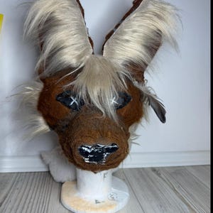 May include: A brown animal mask featuring large, upright ears covered in long, cream-colored fur. The mask has black, diamond-shaped eye patches with white stitched X details, a dark brown snout, and a glossy black nose. The fur texture is shaggy.