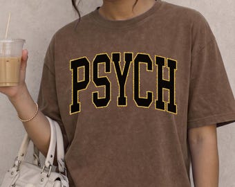 May include: A brown mineral-washed t-shirt featuring the word PSYCH printed in large black block letters with a yellow outline across the chest. The shirt has a crew neckline and short sleeves, presenting a vintage-inspired aesthetic.