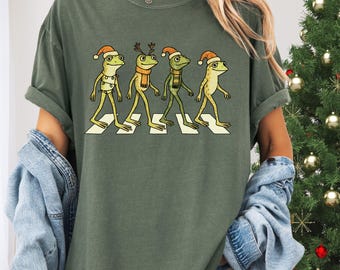 May include: Olive green t-shirt featuring a graphic of four frogs crossing a crosswalk, each wearing a Santa hat and scarf. The shirt has the text "MOSS Comfort Colors C1717" at the bottom left.
