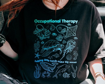 Może przedstawiać: Czarny t-shirt z jasnoniebieskim nadrukiem graficznym przedstawiającym różne stworzenia morskie, w tym ośmiornicę, żółwia morskiego, meduzę, wieloryba i delfina. Tekst głosi Occupational Therapy oraz helping little hands make big waves.