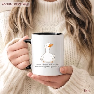 Op de afbeelding: Een witte 325 ml keramische koffiemok met een zwarte handgreep en binnenkant. De mok is voorzien van een cartoonillustratie van een witte eend met een oranje snavel en de tekst: I don't struggle with autism. I'm actually pretty good at it.