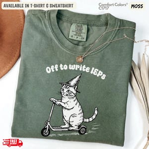Könnte beinhalten: Moosgrünes Comfort Colors Sweatshirt mit dem Text "Off to write IEPs" und einer Grafik einer Katze mit Hexenhut auf einem Roller. Erhältlich als T-Shirt und Sweatshirt.