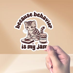 Könnte beinhalten: Ein weißer Aufkleber mit einer Comic-Katze, die Marmelade auf ein Stück Brot streicht. Der Text auf dem Aufkleber lautet "because behavior is my jam". Das Marmeladenglas ist mit "behavior" beschriftet.