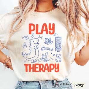 Peut inclure: T-shirt ivoire avec l'inscription "PLAY THERAPY" en rouge. Le motif comprend des dessins bleus de jouets, dont un dinosaure, un avion, un train et un ours en peluche. Fabriqué par Comfort Colors.