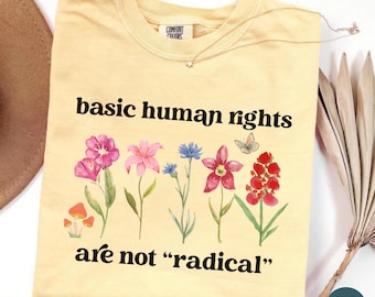May include: A butter yellow t-shirt featuring the black text basic human rights are not radical. The design includes a colorful illustration of various wildflowers, mushrooms, and a small butterfly centered between the two lines of text.