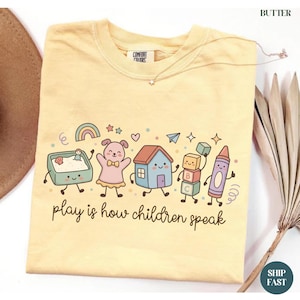 Peut inclure: Un t-shirt jaune beurre avec une impression graphique représentant un bac à sable, un lapin rose, une maison bleue, des blocs alphabet et un crayon violet avec des visages. Le texte "play is how children speak" est imprimé en brun.