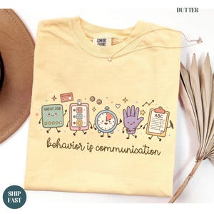 May include: A butter yellow t-shirt featuring a graphic of five anthropomorphic school supplies including a smiley face chart, a tablet, a clock, a hand, and a clipboard. The text "behavior is communication" is printed in brown below the graphic.