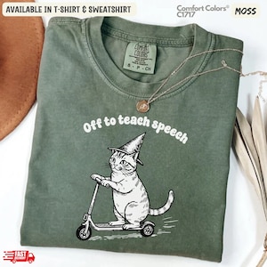 Op de afbeelding: Een mosgroen t-shirt met een zwart-witte afbeelding van een kat met een puntmuts op een step. De tekst boven de kat luidt "Off to teach speech". Het t-shirt is voorzien van een Comfort Colors-merklabel aan de binnenzijde van de hals.