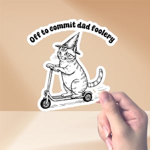 Op de afbeelding: Een zwart-witte sticker met een kat met een tovenaars hoed op een scooter. De tekst "Off to commit dad foolery" staat boven de kat. De sticker heeft een witte rand.