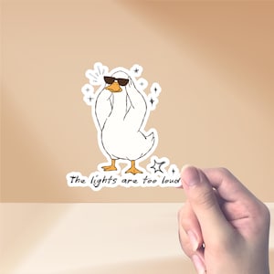 May include: A white sticker with a cartoon duck wearing sunglasses and covering its eyes. The duck is surrounded by star-shaped sparkles. The text "The lights are too loud" is below the duck.