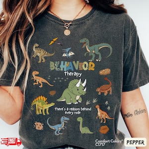 Può includere: T-shirt grigio scuro con grafica di dinosauri colorati. La maglietta presenta le parole "BEHAVIOR therapy" e "there's a reason behind every roar". La maglietta è una Comfort Colors C1717 in Pepper.