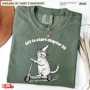 May include: Moss green t-shirt with the text "Off to start chapter 30" and a graphic of a cat wearing a witch hat riding a scooter. The shirt is available in T-shirt and sweatshirt styles.