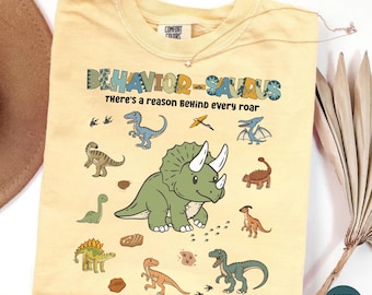 May include: A butter yellow t-shirt featuring a central green triceratops illustration surrounded by various smaller dinosaur graphics. The text reads "BEHAVIOR-SAURUS" and "There's a reason BeHind every roar" in a multi-colored, playful font.