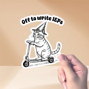 Könnte beinhalten: Ein schwarz-weißer Aufkleber mit einer Katze mit Hexenhut, die einen Roller fährt. Der Text "Off to write IEPs" steht über der Katze. Der Aufkleber wird von einer Hand gehalten.