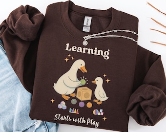 May include: Dark chocolate-colored sweatshirt with the text "Learning Starts with Play." The graphic features two ducks, a cube, and various toys. A silver necklace is draped over the sweatshirt.