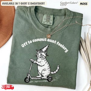 Puede incluir: Camiseta verde musgo con el texto "Off to commit aunt foolery" y un gráfico de un gato con sombrero de bruja en un patinete. Disponible en camiseta y sudadera.