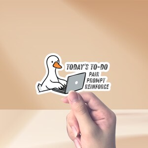 Könnte beinhalten: Ein weißer Aufkleber mit einer Cartoon-Gans, die an einem Laptop arbeitet. Die Gans hat einen orangefarbenen Schnabel und sitzt vor einem grauen Laptop. Der Text auf dem Aufkleber lautet "TODAY'S TO-DO, PAIR, PROMPT, REINFORCE."