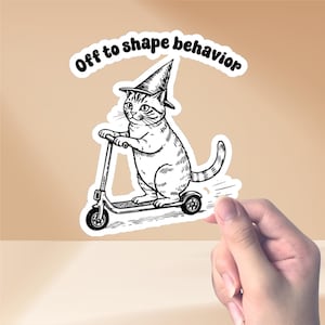 May include: A black and white sticker featuring a cat wearing a witch hat and riding a scooter. The sticker has the text "Off to shape behavior" in a white font.