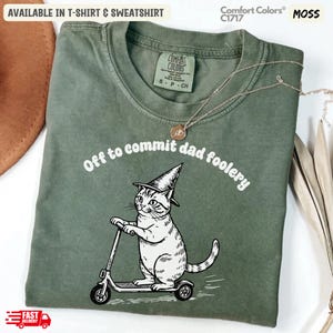 Op de afbeelding: Mosgroen T-shirt met de tekst "Off to commit dad foolery" boven een cartoonkat op een scooter met een tovenaars hoed. Verkrijgbaar in t-shirt en sweatshirt. Comfort Colors C1717.
