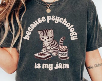 May include: A dark grey t-shirt featuring a cute cartoon cat spreading jam on a slice of bread. The text "because psychology is my jam" is printed on the shirt.