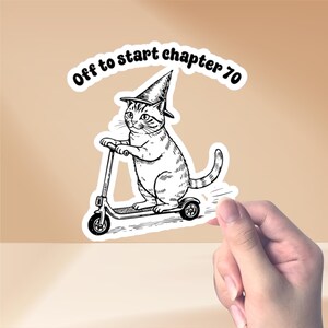 May include: Black and white sticker featuring a cat riding a scooter and wearing a witch hat. The text "Off to start chapter 70" is arched above the cat. The sticker is held by a hand.