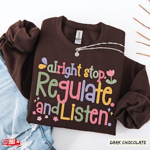 Op de afbeelding: Donker chocoladekleurige sweatshirt met de tekst "Alright stop. Regulate and Listen" in kleurrijke letters. Een zilveren ketting met een zonnehanger ligt op de sweatshirt. De sweatshirt ligt naast een spijkerbroek.