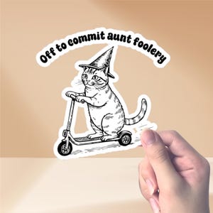 Puede incluir: Una pegatina en blanco y negro que muestra un gato montado en un patinete, con un sombrero de bruja y el texto "Off to commit aunt foolery" en la parte superior. La pegatina está sujeta por una mano.