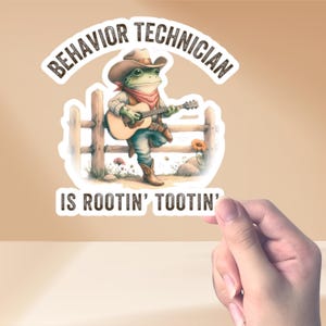 May include: A sticker featuring a cartoon frog wearing a cowboy hat and playing a guitar. The text on the sticker reads "Behavior Technician is rootin' tootin'".